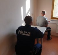 Na zdjęciu policjant siedzący przy stoliku oraz odwrócony tyłem zatrzymany mężczyzna , który był poszukiwany przez Policjię.