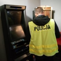 Na zdjęciu policjant w cywilu ubrany w kamizelkę odblaskową z napisem POLICJA wykonujący czynności w nielegalnym salonie gier.