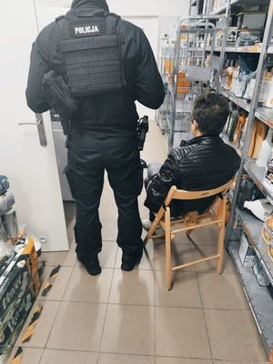 na zdjęciu widoczny jest umundurowany policjant , obok na krześle siedzi osoba zatrzymana z kajdankami na nadgarstkach , które ma zapięte do tyłu